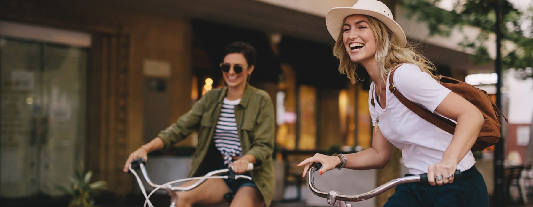 two young women smile as they ride bicycles down a city sidewalk
