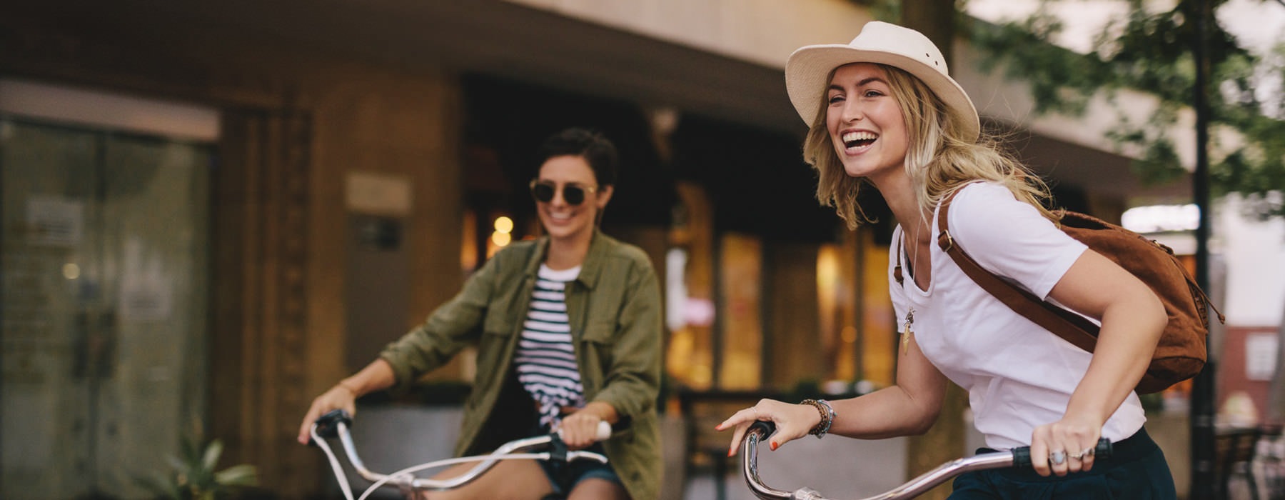 two young women smile as they ride bicycles down a city sidewalk